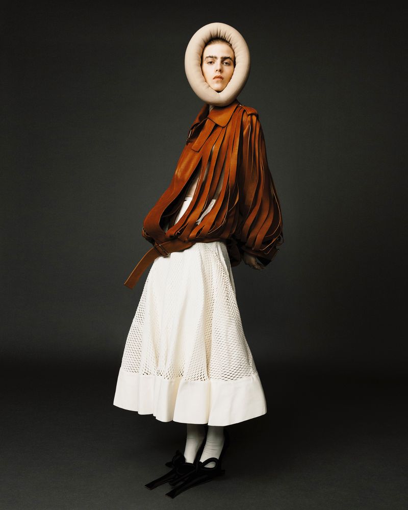 A fashion portrait shows a woman in a tan and brown leather jacket and white skirt, topped with a padded ring-shaped headpiece on a grey background.