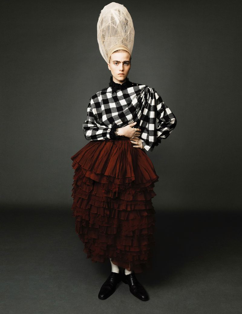 Fashion model poses in black and white checkered top, layered red skirt, and tall white wig in studio against gray backdrop.