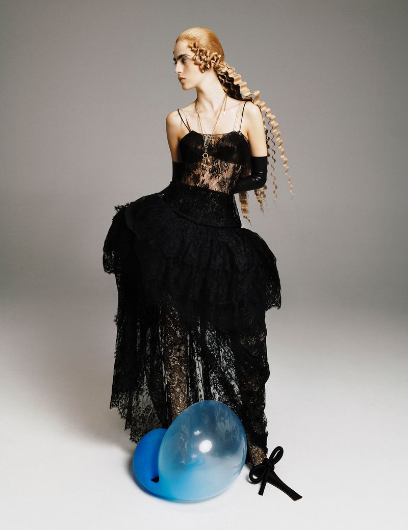 A slender fashion model in a striking black lace gown poses in a photo studio, with her long blonde hair braided and arranged.
