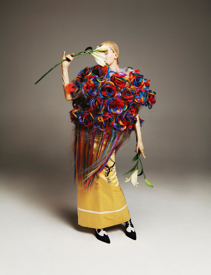 Adult fashion model in yellow dress and floral cape poses with white lilies in a studio setting with neutral gray background.