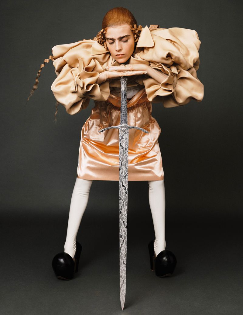 Portrait of a person in an avant-garde dress and heels holding a sword. Dark studio backdrop with dramatic lighting.  Fashion editorial.
