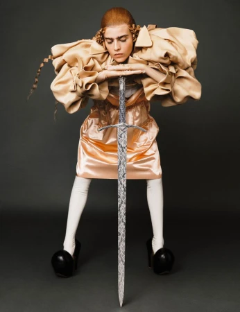 Portrait of a person in an avant-garde dress and heels holding a sword. Dark studio backdrop with dramatic lighting.  Fashion editorial.