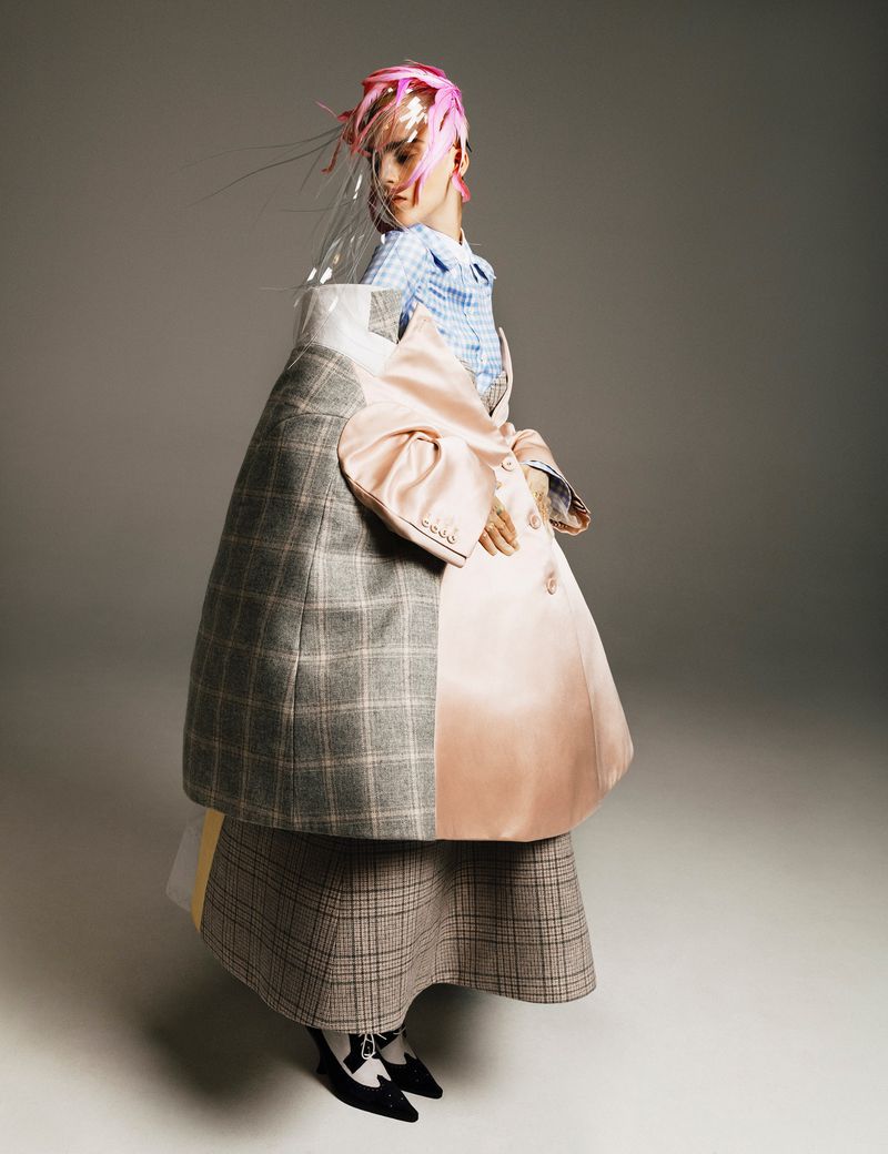 Woman in a unique high-fashion editorial studio shot, wearing a pink and grey oversized coat and skirt, with a pink hair accessory.
