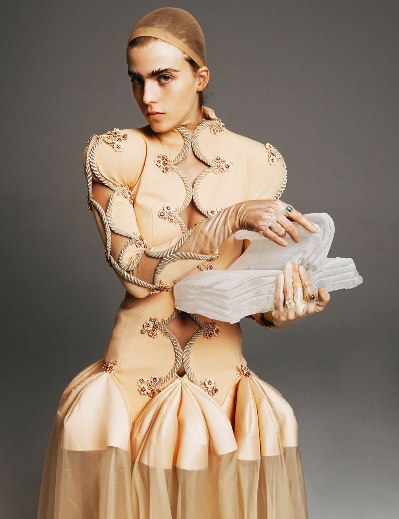 A woman in an embellished, light-orange couture dress holds a translucent object in a studio setting, gazing intensely at the viewer.