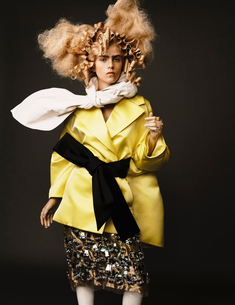 Portrait of a fashion model in a yellow satin coat with a large black bow and a curly blond hairstyle against a grey background.