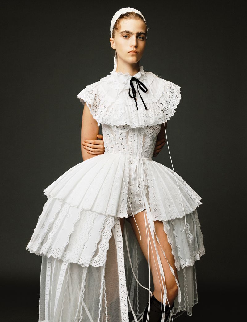 Elegant portrait of an adult female fashion model wearing a white dress with ruffles and a head covering against a gray background.