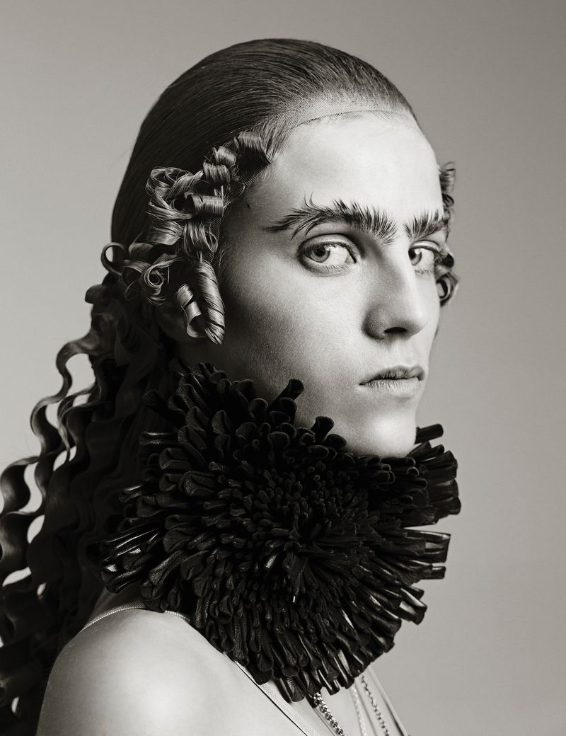 Close-up portrait of a person with an elaborate hairstyle, dark ruffled collar, and serious expression in a studio environment.