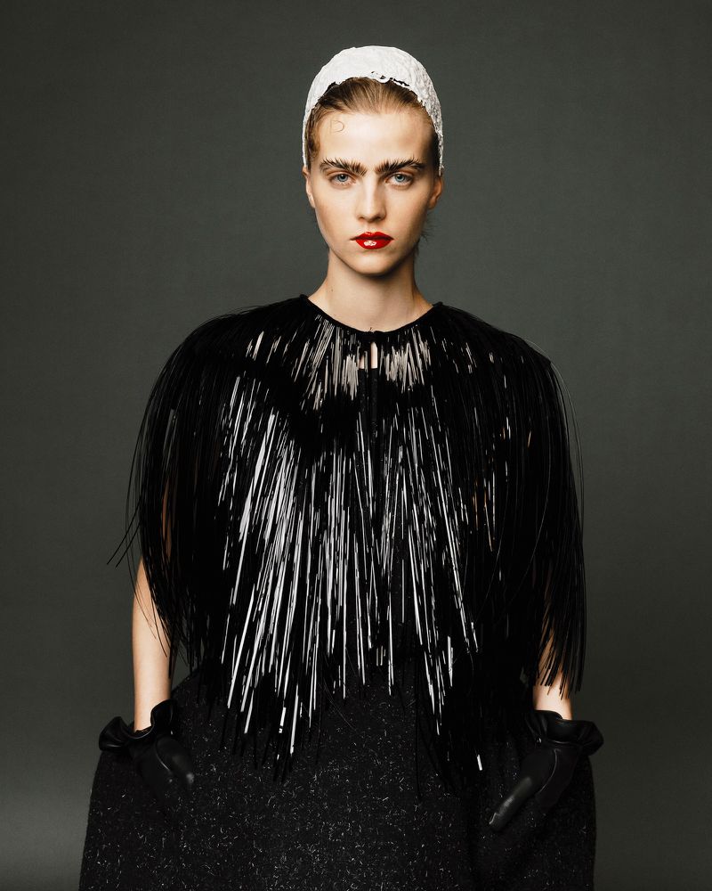 Adult female fashion model poses in a glossy black structural cape, white head covering, dark dress, and gloves against a gray studio backdrop.