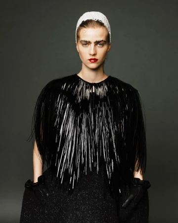 Adult female fashion model poses in a glossy black structural cape, white head covering, dark dress, and gloves against a gray studio backdrop.