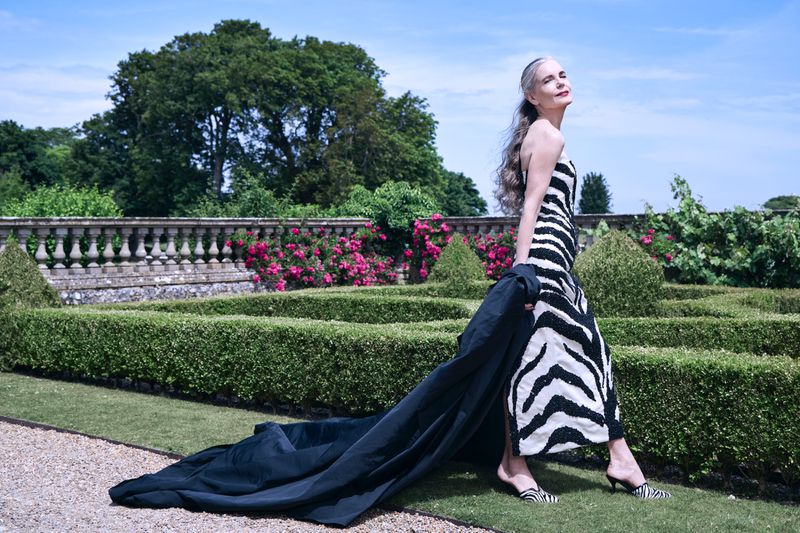 Adult female fashion model in a zebra-striped dress poses in a sunny manicured garden with green hedges, a stone wall, and pink flowers.
