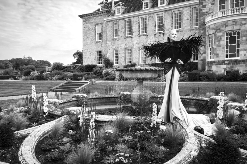 Woman in dramatic black feathered dress stands in an ornate garden with a fountain. A historic stone building is in the background.