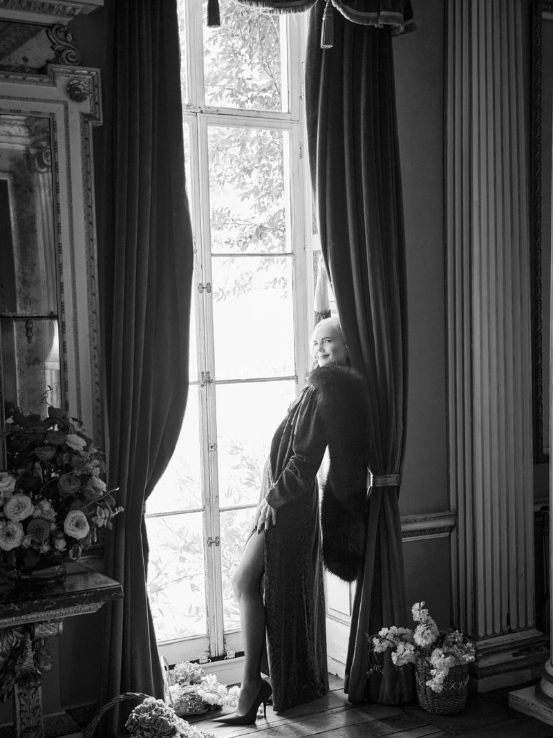 Elegant blonde woman in long gown poses by large window with dramatic curtains and flower arrangements in a stately room, black and white.