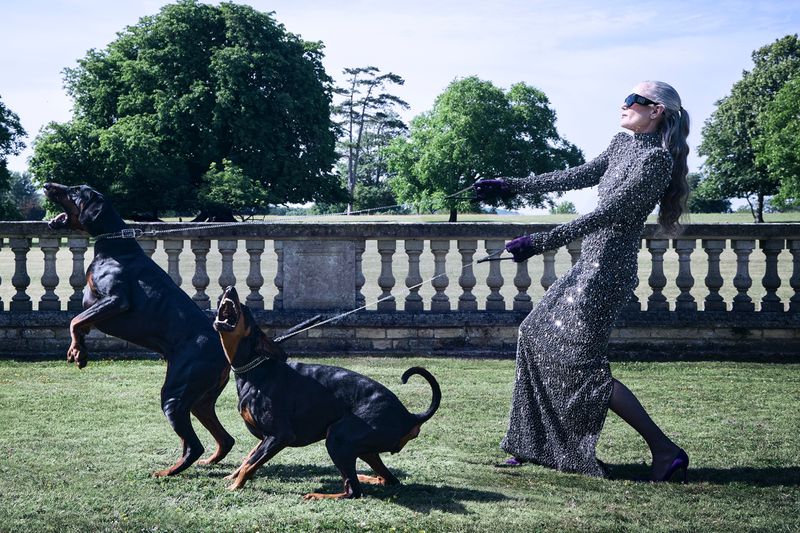Elegant mature woman in sparkling gown holds back two Doberman dogs in a park setting with a stone wall and large tree backdrop.