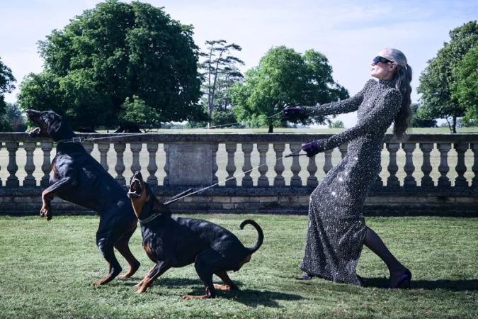 Elizabeth McGovern in a sparkling evening gown holding leashes to two barking Dobermans on a green lawn.