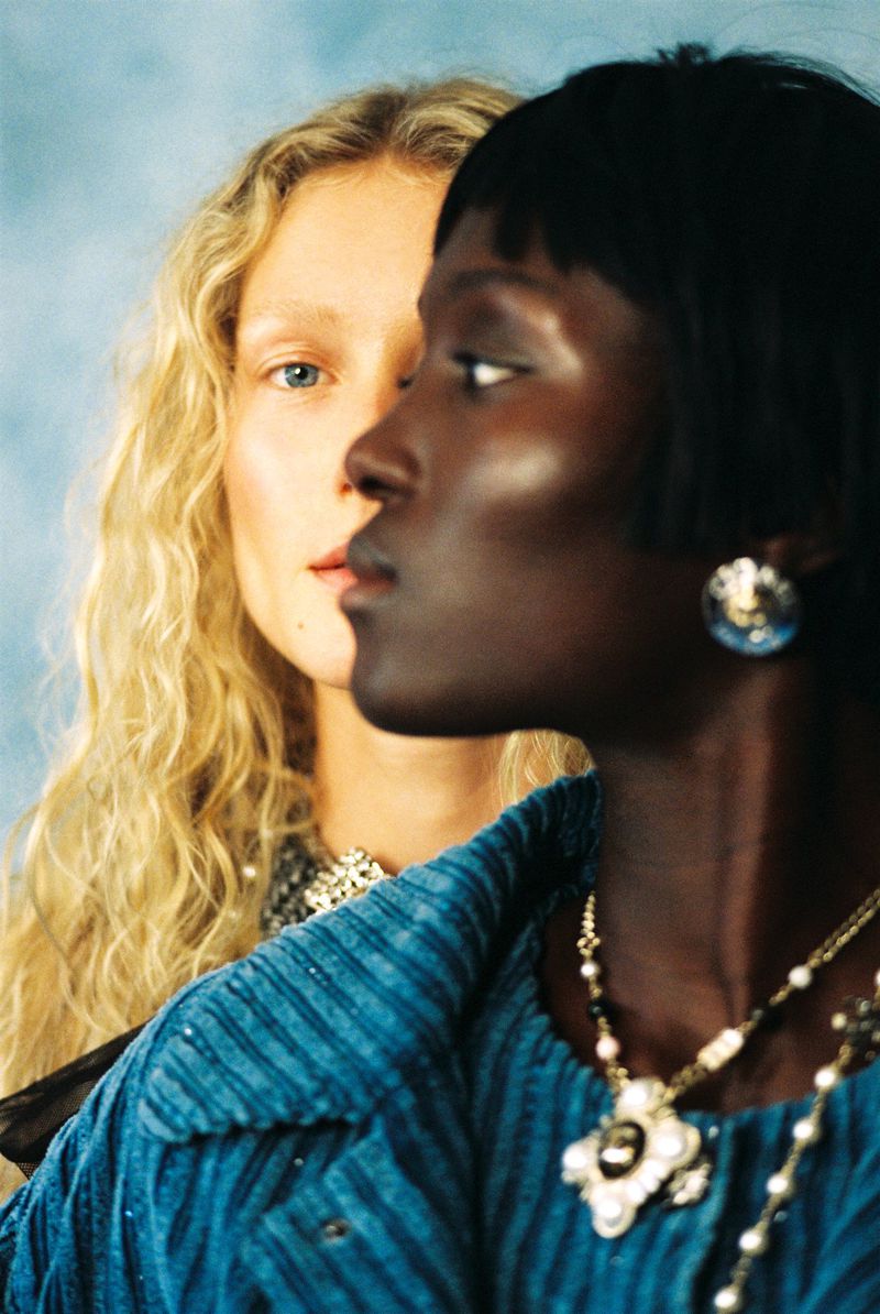 Close-up profile of a woman in a blue corduroy jacket and ornate pendant necklace alongside a blonde model against a blue backdrop.