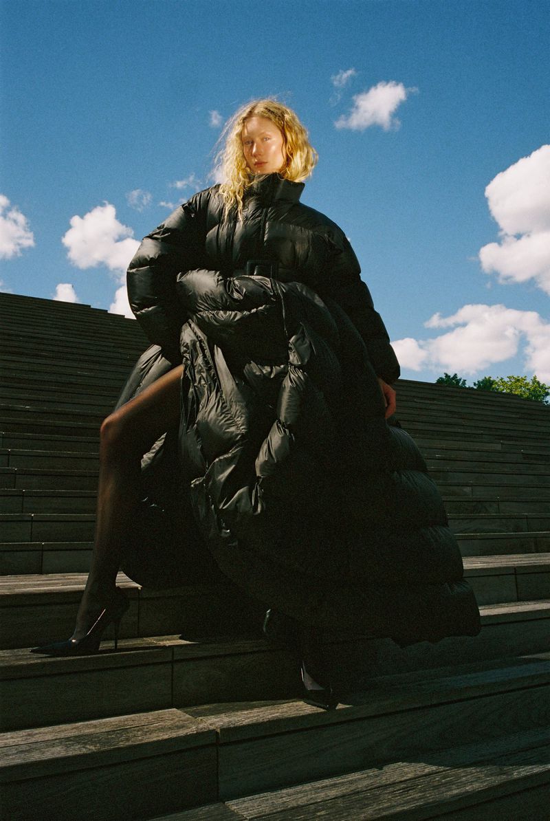 Woman in black puffer coat and heels poses on concrete steps under a bright blue sky with fluffy white clouds in an outdoor fashion shoot.