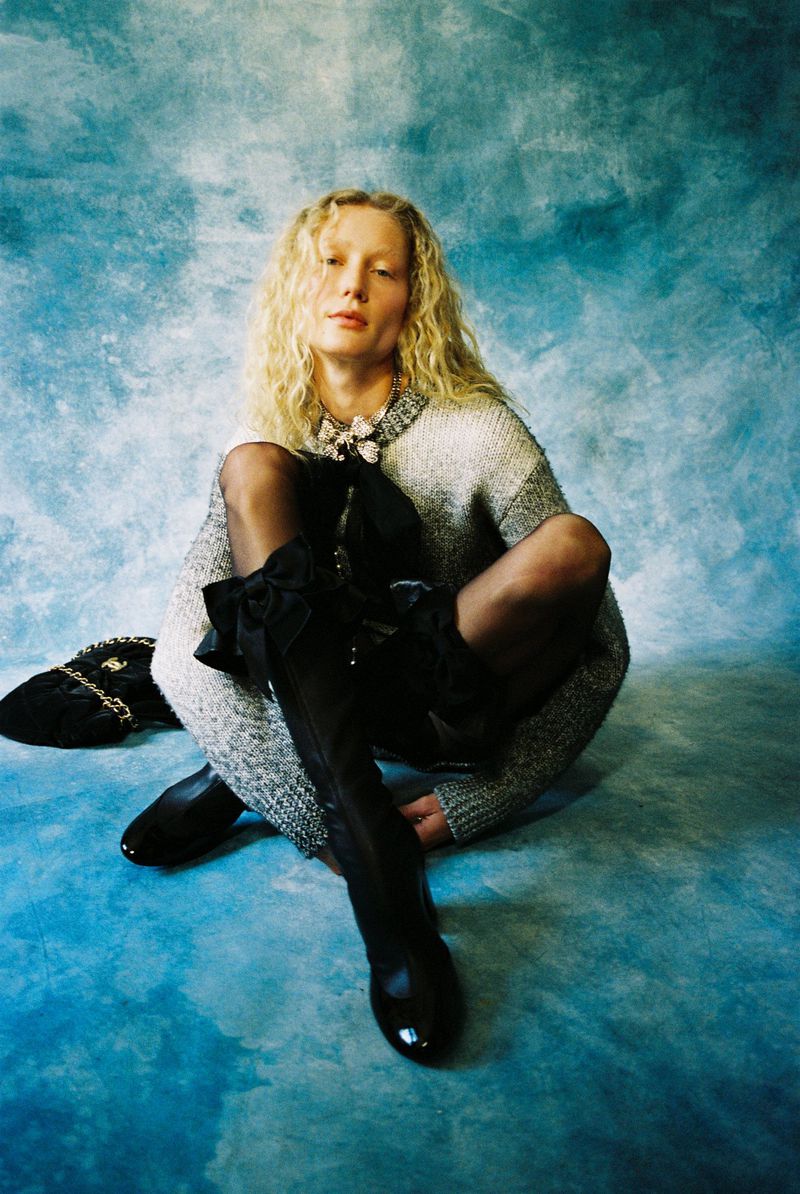 Blonde woman sits cross-legged wearing thigh-high boots with black bows, a gray sweater, and a pearl necklace against a blue background.