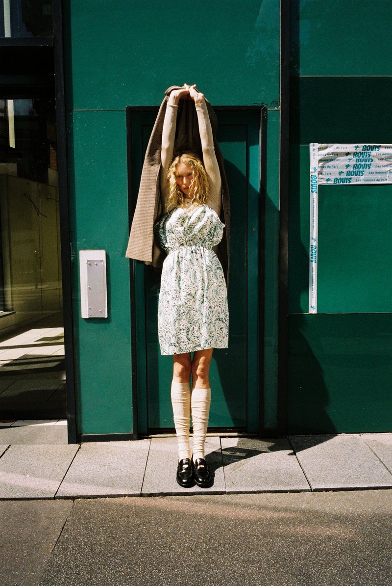 Woman in floral dress and knee socks stands against green wall, holding a tan coat overhead on a city street, natural light.