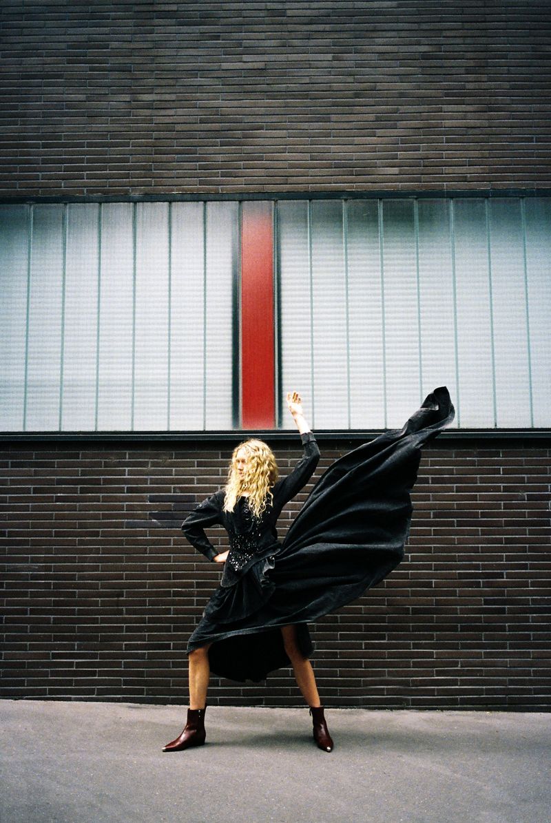 Woman in black dress poses with arms raised outdoors against brick wall and window, fashion editorial style with movement.