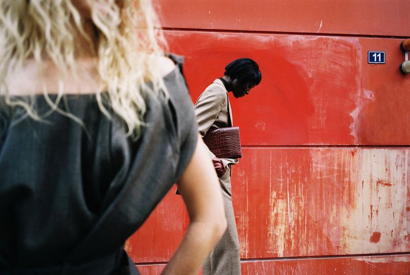 Two women in suits near a red wall. One woman has blond curly hair and faces away, the other has an auburn bag. 