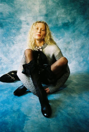 Model Sandra Martens crouched in a grey knit sweater and black boots against a mottled blue painted backdrop.