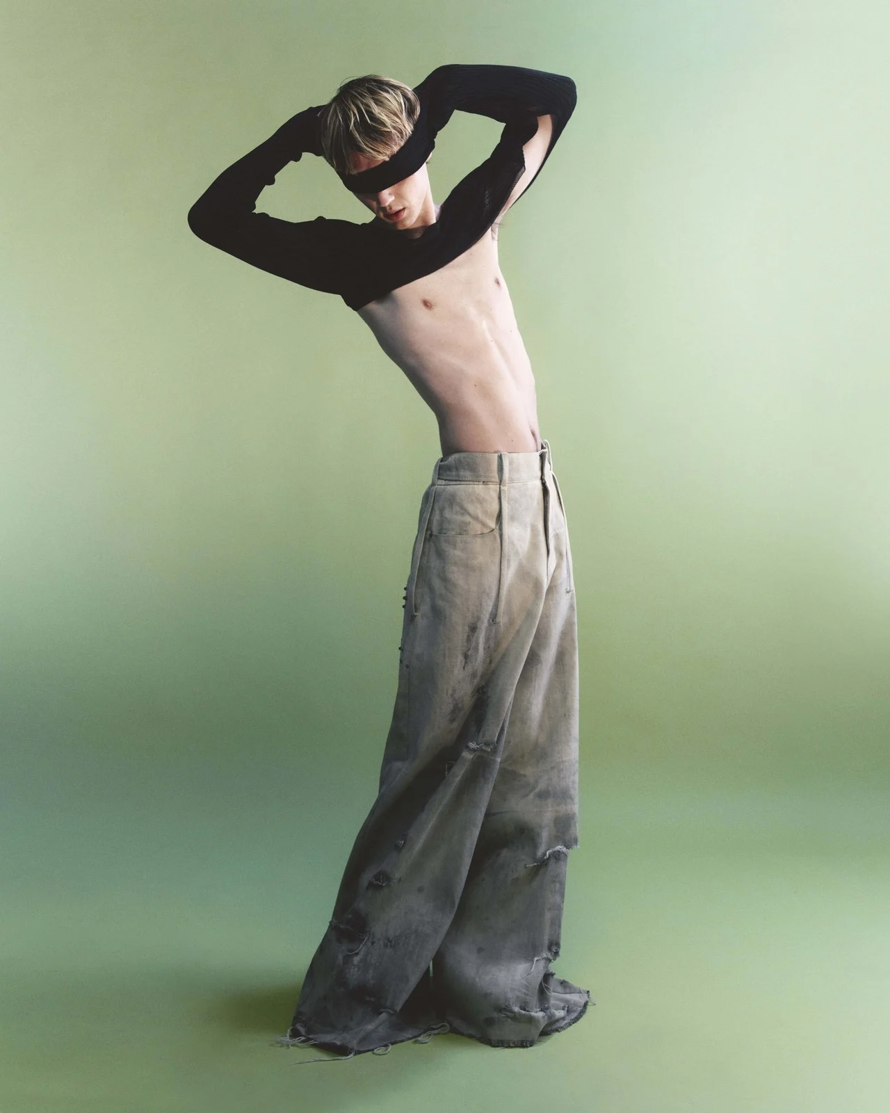 Male fashion model poses shirtless in ripped jeans and black sleeves in a full shot against a light-green seamless backdrop.