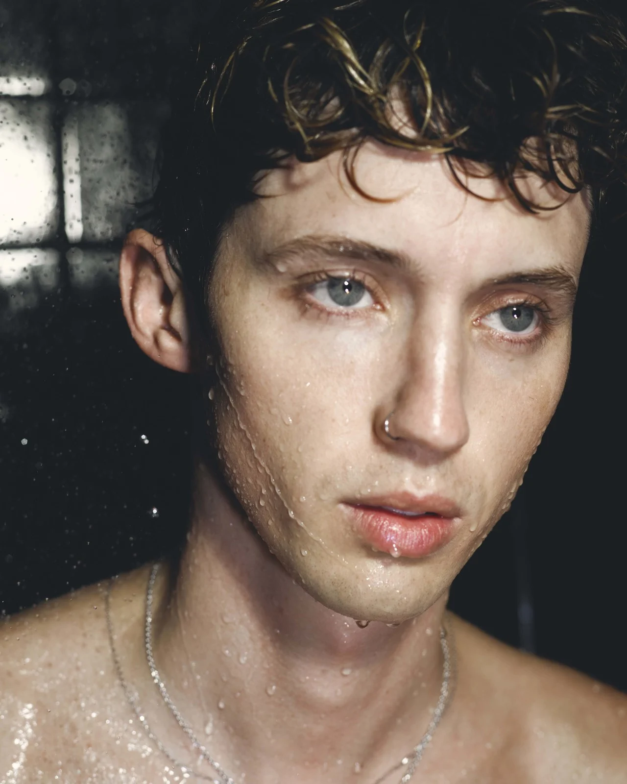 Close-up portrait of a young adult male with curly hair and water droplets on his face, set against a dark, blurred background.