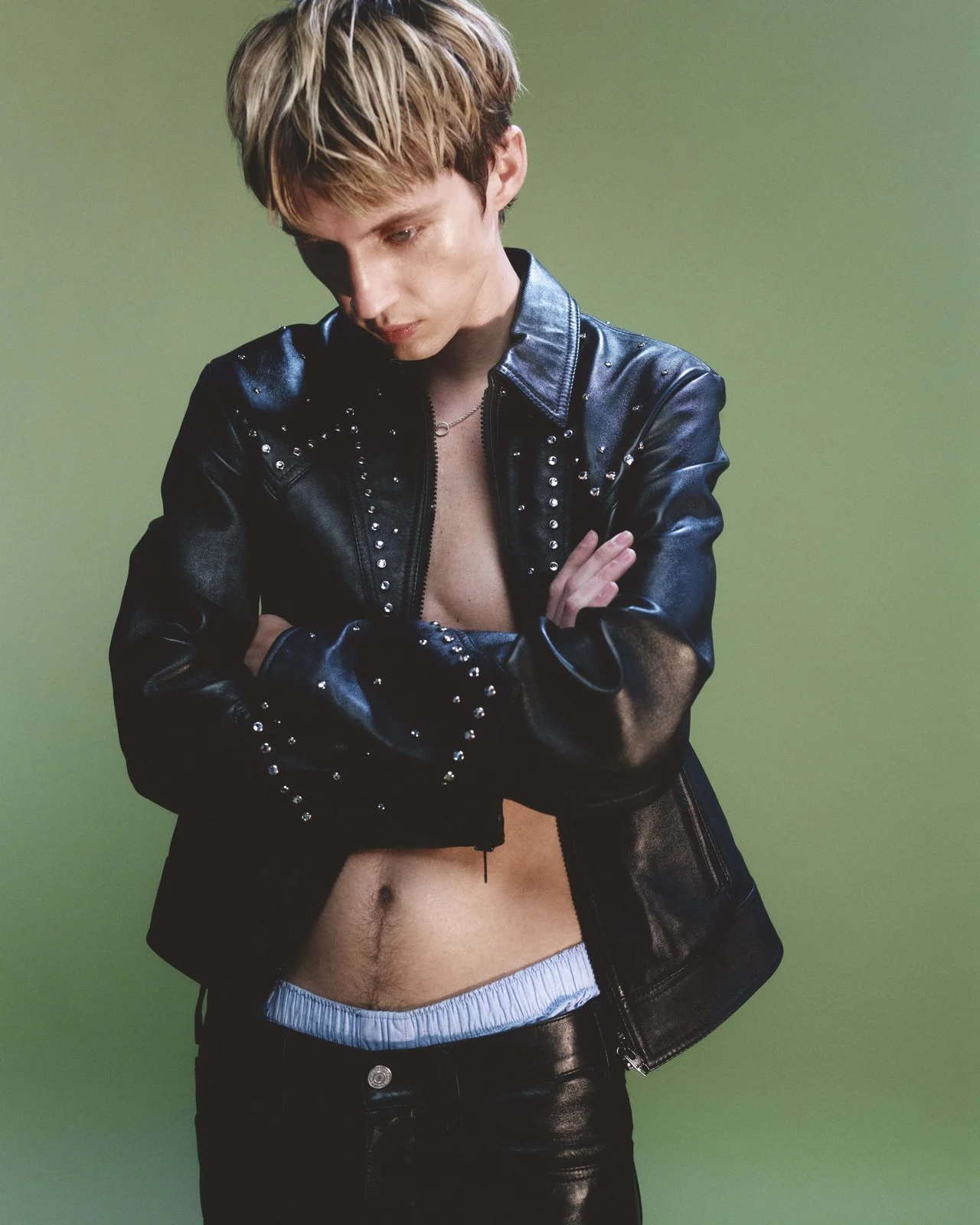 Fashionable young adult poses indoors wearing an open black leather jacket, black pants, and light blue boxers against a green backdrop.