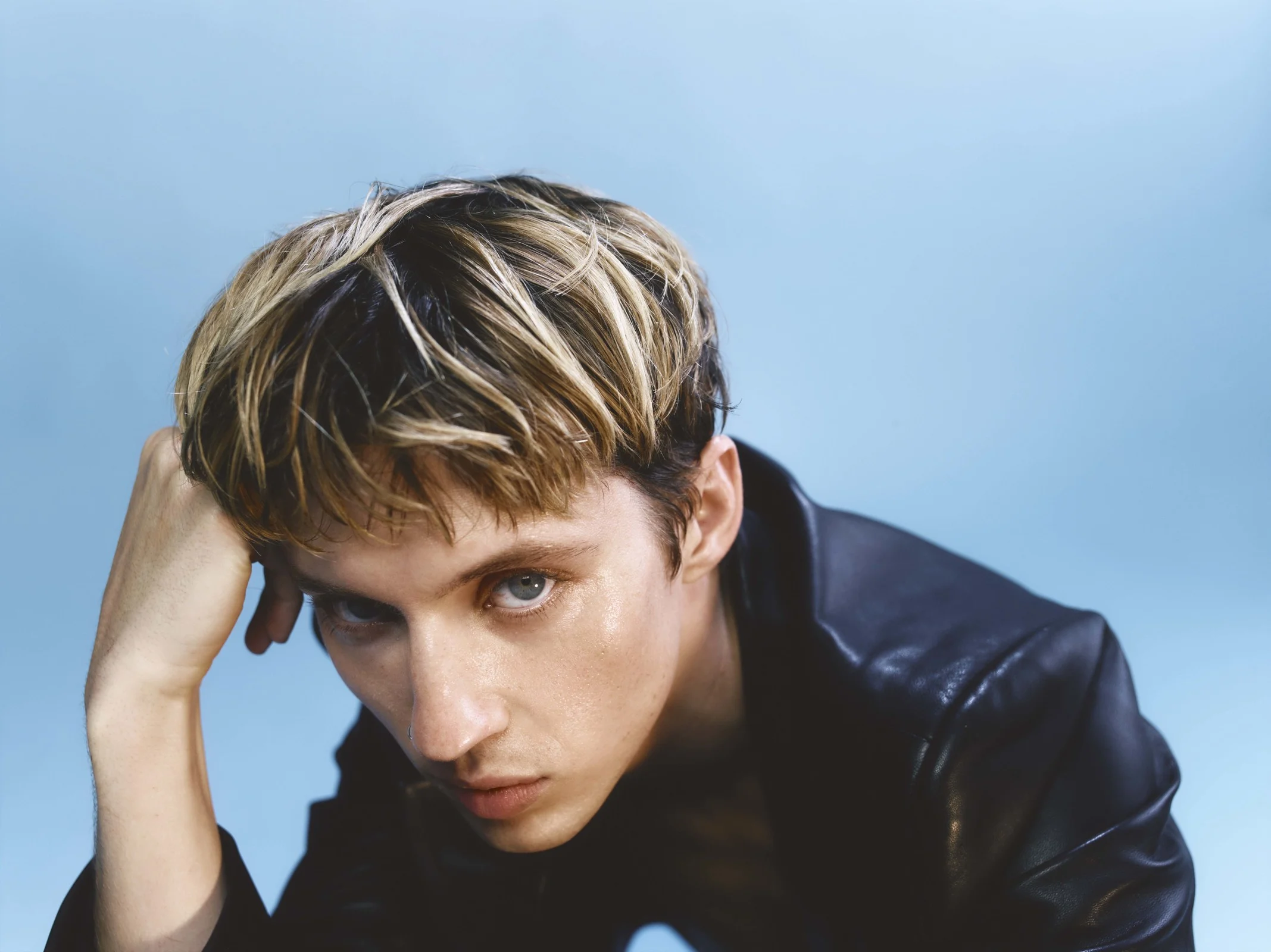 Close-up portrait of Troye Sivan wearing a black leather jacket against a light blue background, resting head on hand.