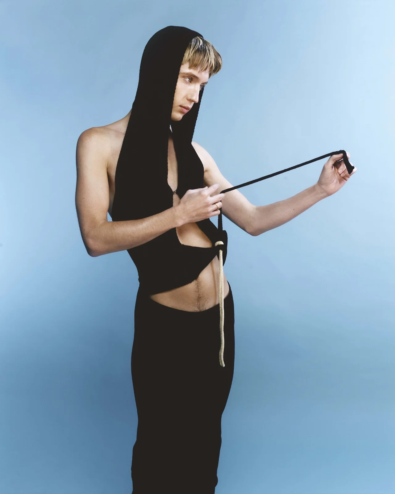 Man poses in black knit halter top with hood and long black knit skirt in front of a light blue studio backdrop in a fashion setting.