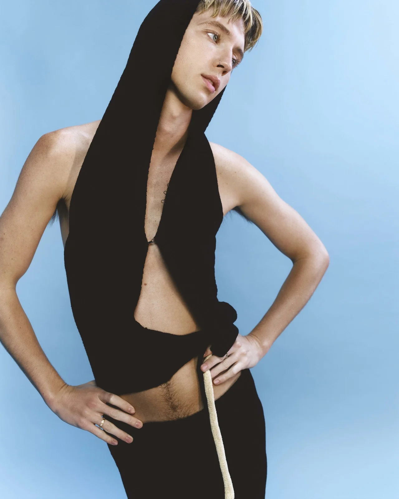 Young adult with blond hair poses in a hooded knit top and pants against a light blue background in a fashion photography studio shot.