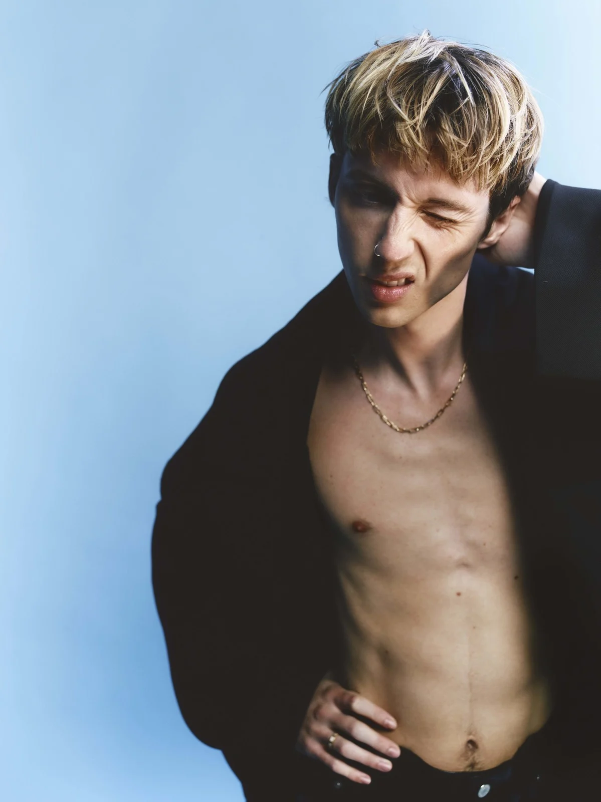 Portrait of a shirtless adult male with light hair, wearing a dark jacket and gold chain necklace, posing against a light blue background.