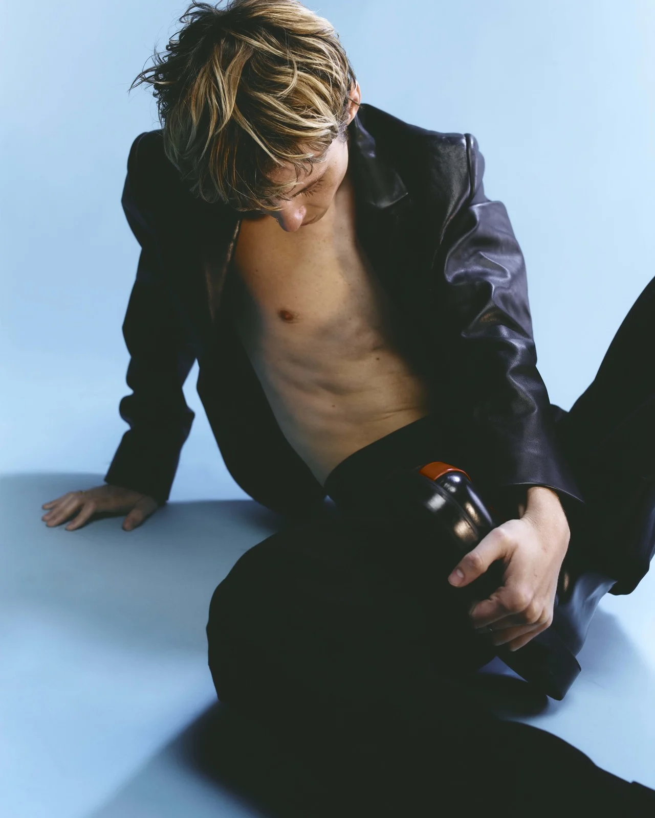 Blonde adult male sits against a light blue backdrop, wearing a black leather jacket and black pants. He is holding a black bottle.