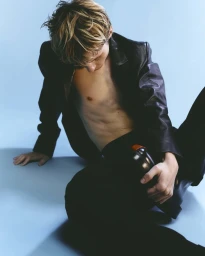 Young blond man wearing a black leather jacket and trousers seated against a blue seamless background