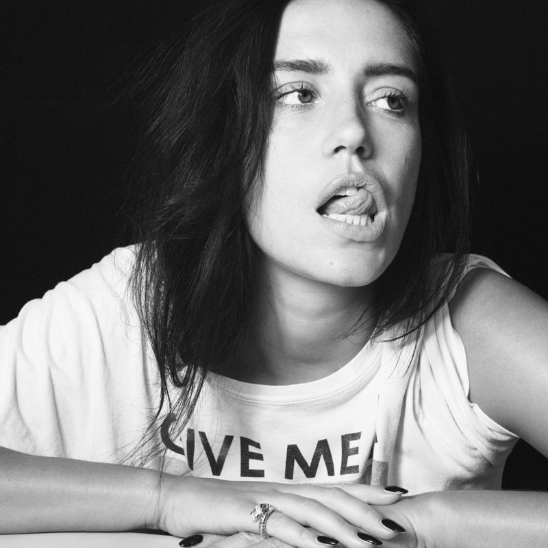Portrait of a young adult female with dark hair sticking her tongue out, wearing a white t-shirt in front of a black background.