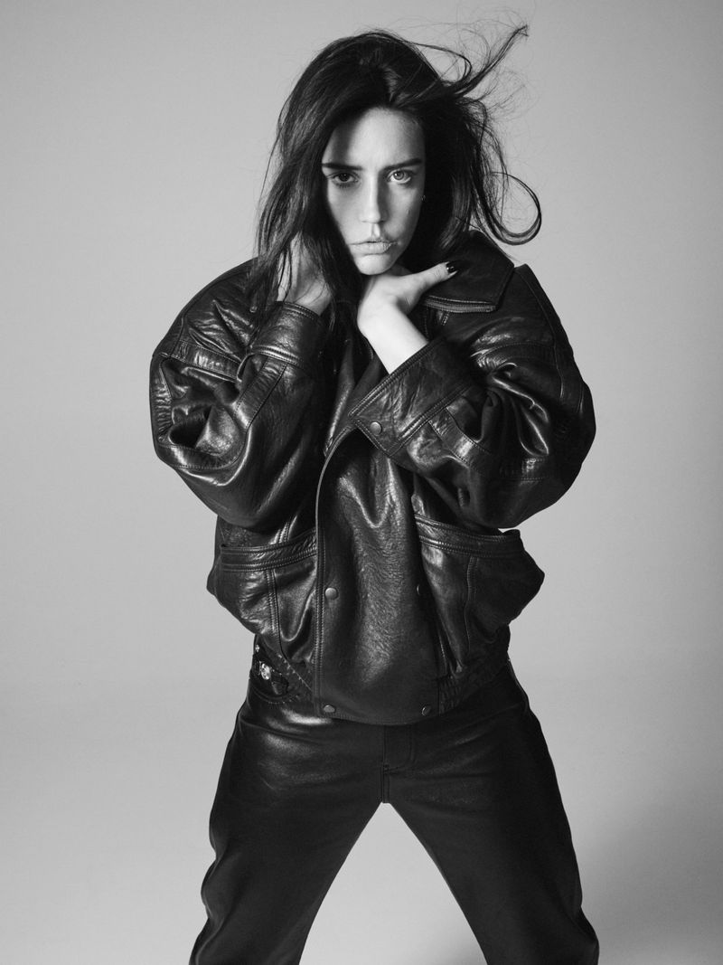 Black and white portrait of a young adult woman with dark hair, wearing a leather jacket and pants in a photo studio with a plain backdrop.
