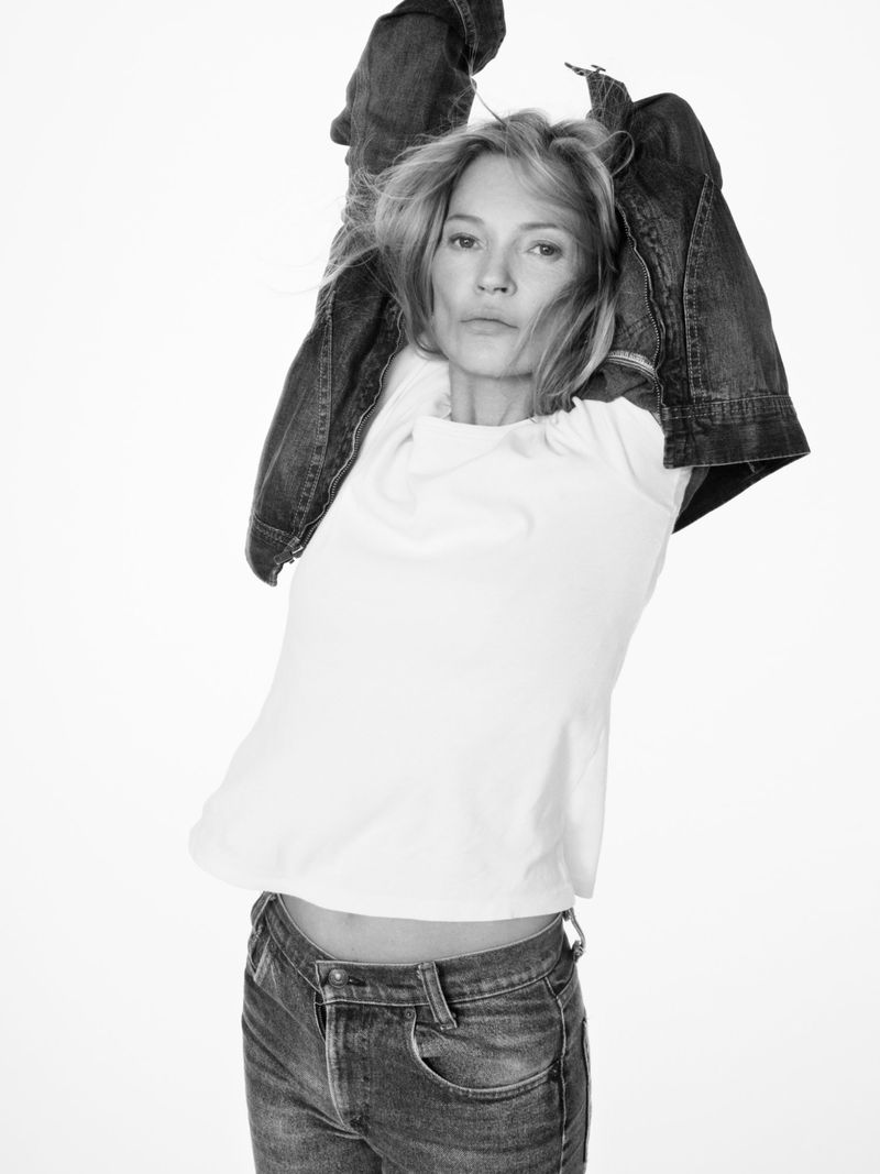 A woman in a white top, blue jeans, and a denim jacket poses against a white backdrop in a black and white fashion photograph.