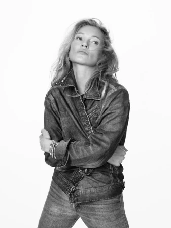 Kate Moss posing in a belted denim jacket and matching jeans with crossed arms against a seamless white studio background.