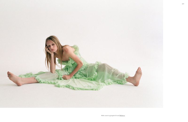 A young woman in a light green ruffled dress reclines on a white floor in a studio setting, her head resting on her hand.