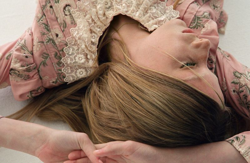 Woman lying down wearing a pink floral top with lace collar. Head angled upward. Long brown hair, fair skin. White background.