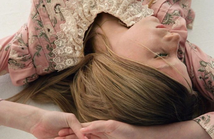 Woman lying down wearing a pink floral top with lace collar. Head angled upward. Long brown hair, fair skin. White background.