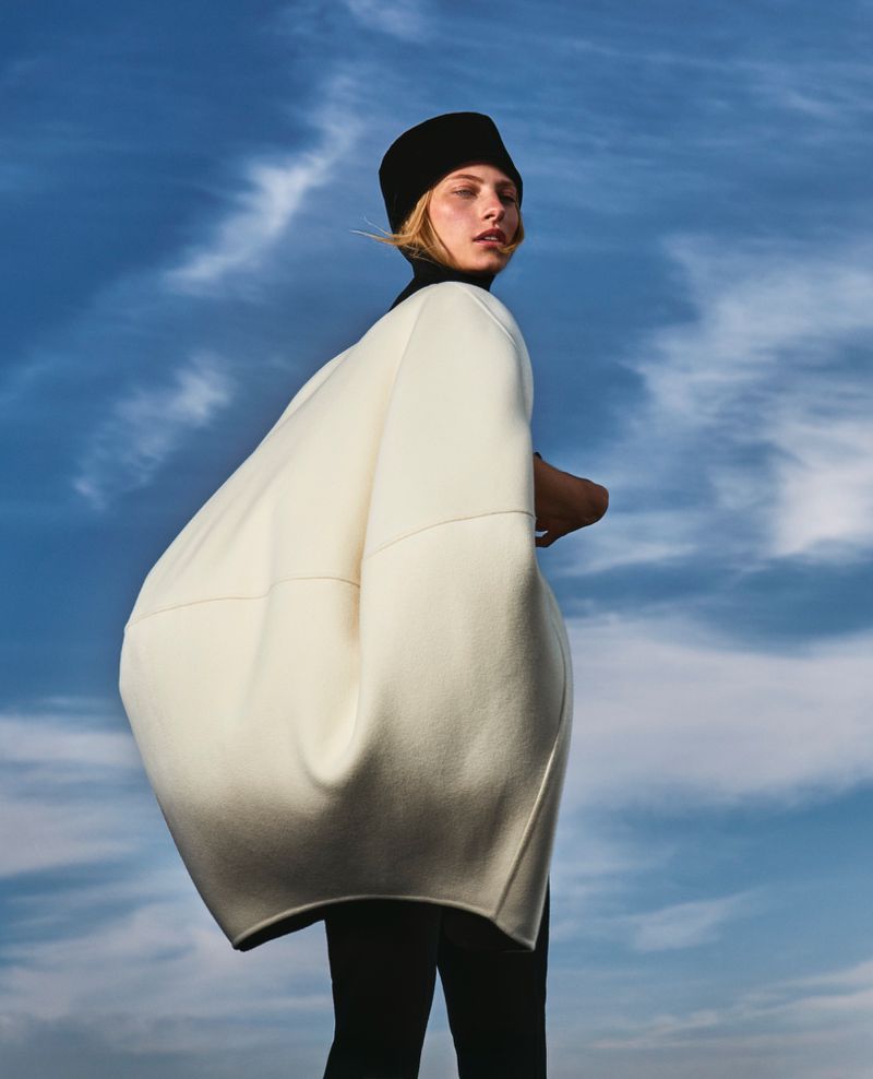 Model Annie Arnander wearing a sculptural off-white voluminous coat and black hat against a blue sky.