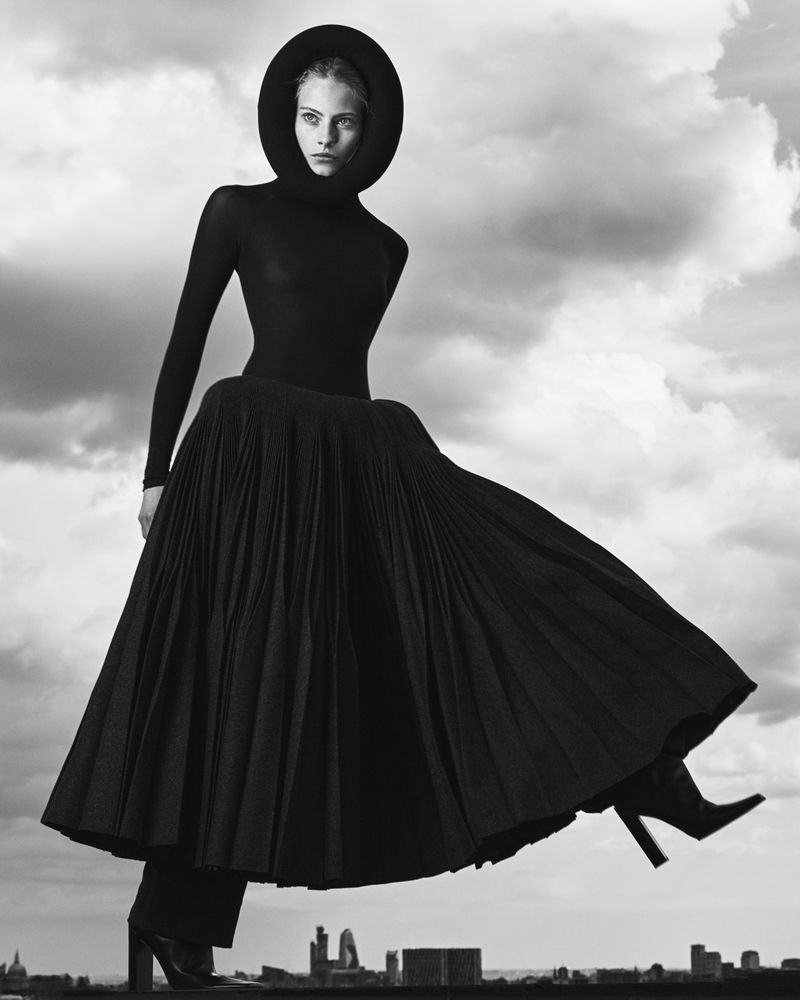 Woman in black formalwear against a cloudy sky and city buildings. Pleated skirt, hood, and high heels in a studio fashion shot.