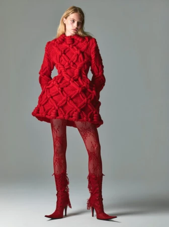 Fashion shot of a fair-skinned woman wearing a bright red knit dress with matching lace tights and high-heeled boots against a grey backdrop.