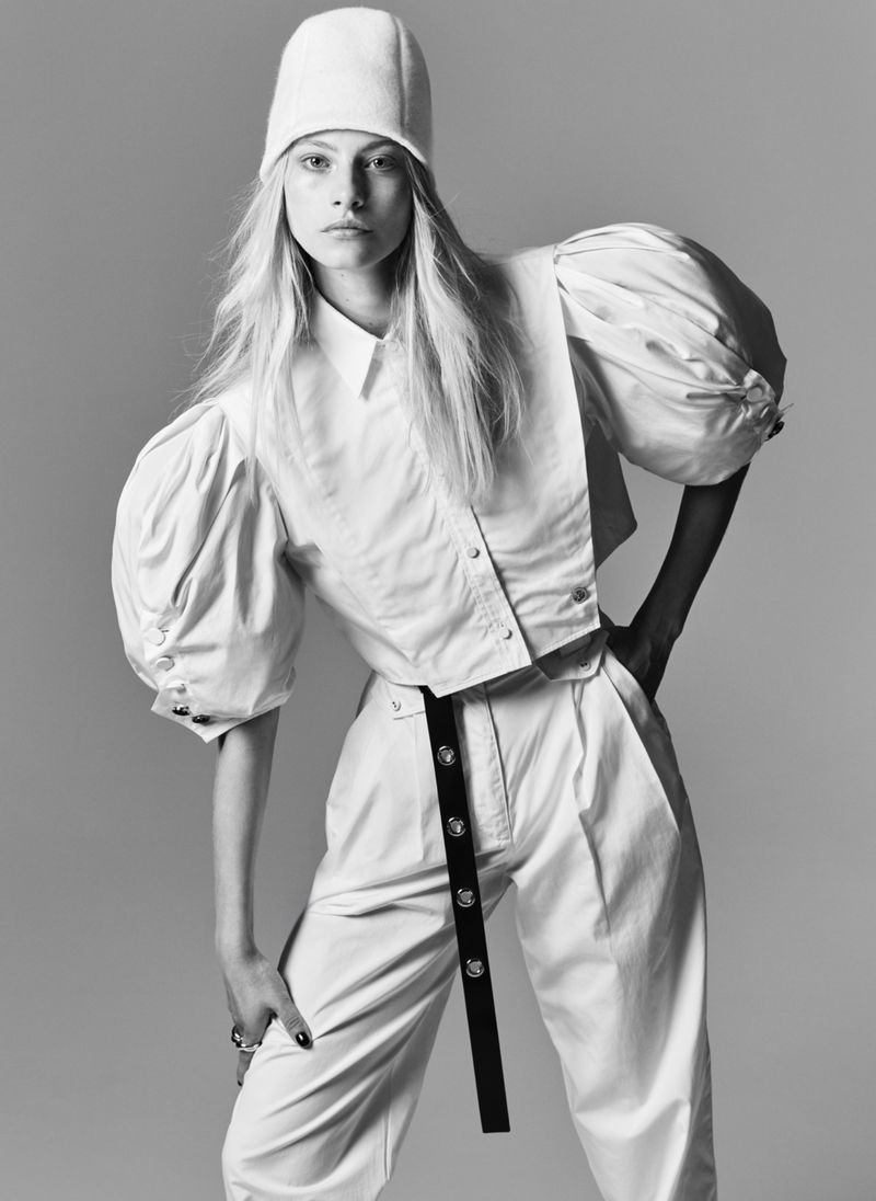 Monochrome fashion shot of a woman in a white hat, collared shirt with puff sleeves, and pants with a dark belt against a grey background.