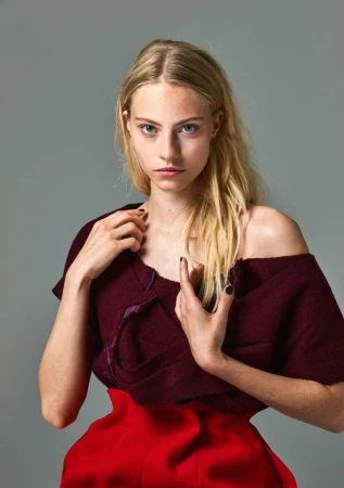 Close-up fashion shot of a fair-skinned young woman with long blonde hair, posing in a red skirt and maroon top against a grey background.