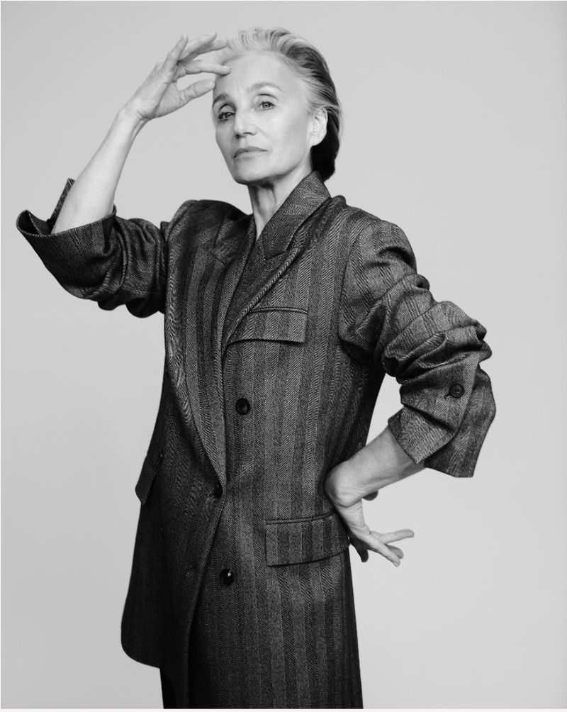Elegant woman in a striped blazer poses in a studio, her hand near her head, against a plain backdrop in a black and white photograph.