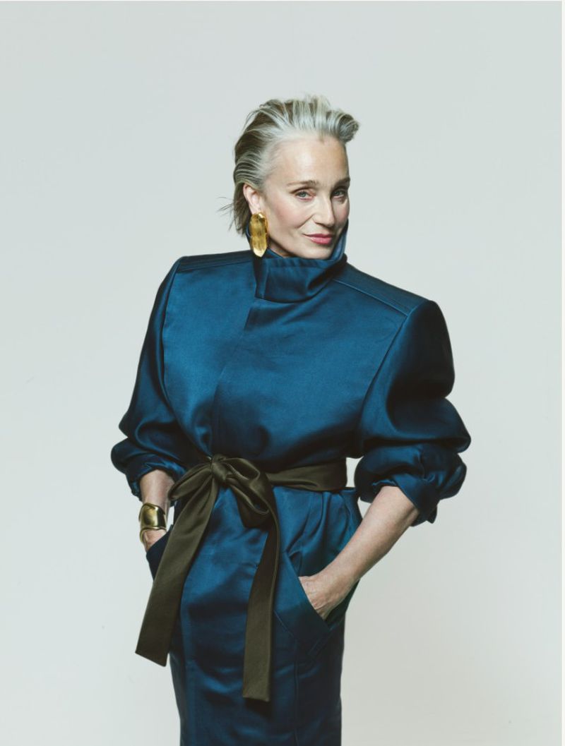 Elegant silver-haired mature woman in blue dress with green belt and gold jewelry posing in a studio portrait, fashion forward.
