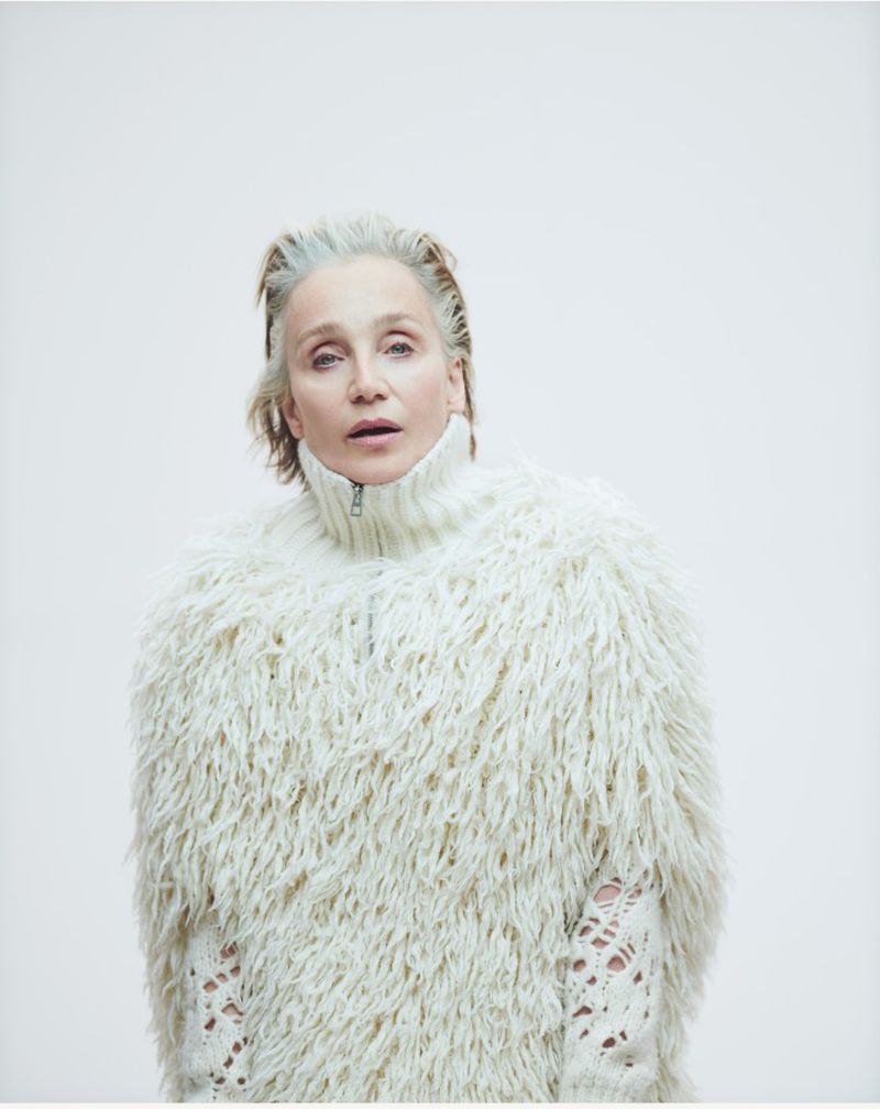Elderly woman in a white, furry sweater stands against a white background, photographed in a fashion-editorial style, capturing texture and mood.