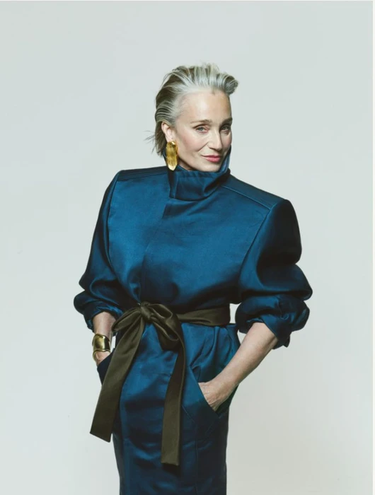 Kristin Scott Thomas in a structured teal coat with a dark tie belt and sculptural gold earrings against a light studio backdrop.